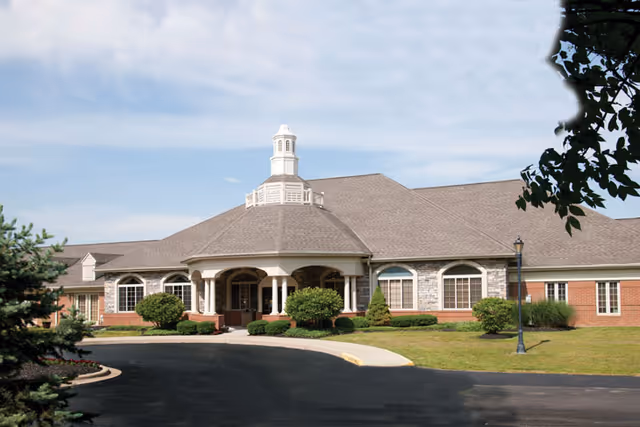 Front exterior view of a single-story senior living facility building with a large covered entrance supported by columns, surrounded by neatly trimmed bushes and a curved driveway. The building has a gray shingled roof with a cupola on top and brick and stone facade under a partly cloudy sky.