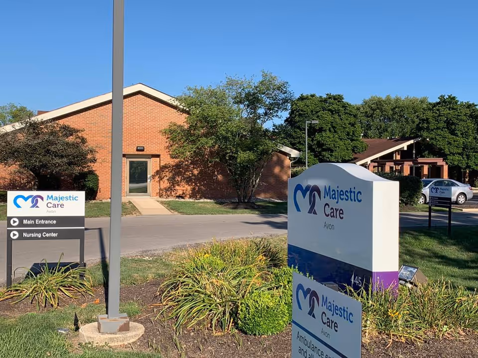 Exterior view of Majestic Care of Avon facility showing brick buildings, green trees, and multiple signs indicating the main entrance, nursing center, and ambulance entrance under a clear blue sky.