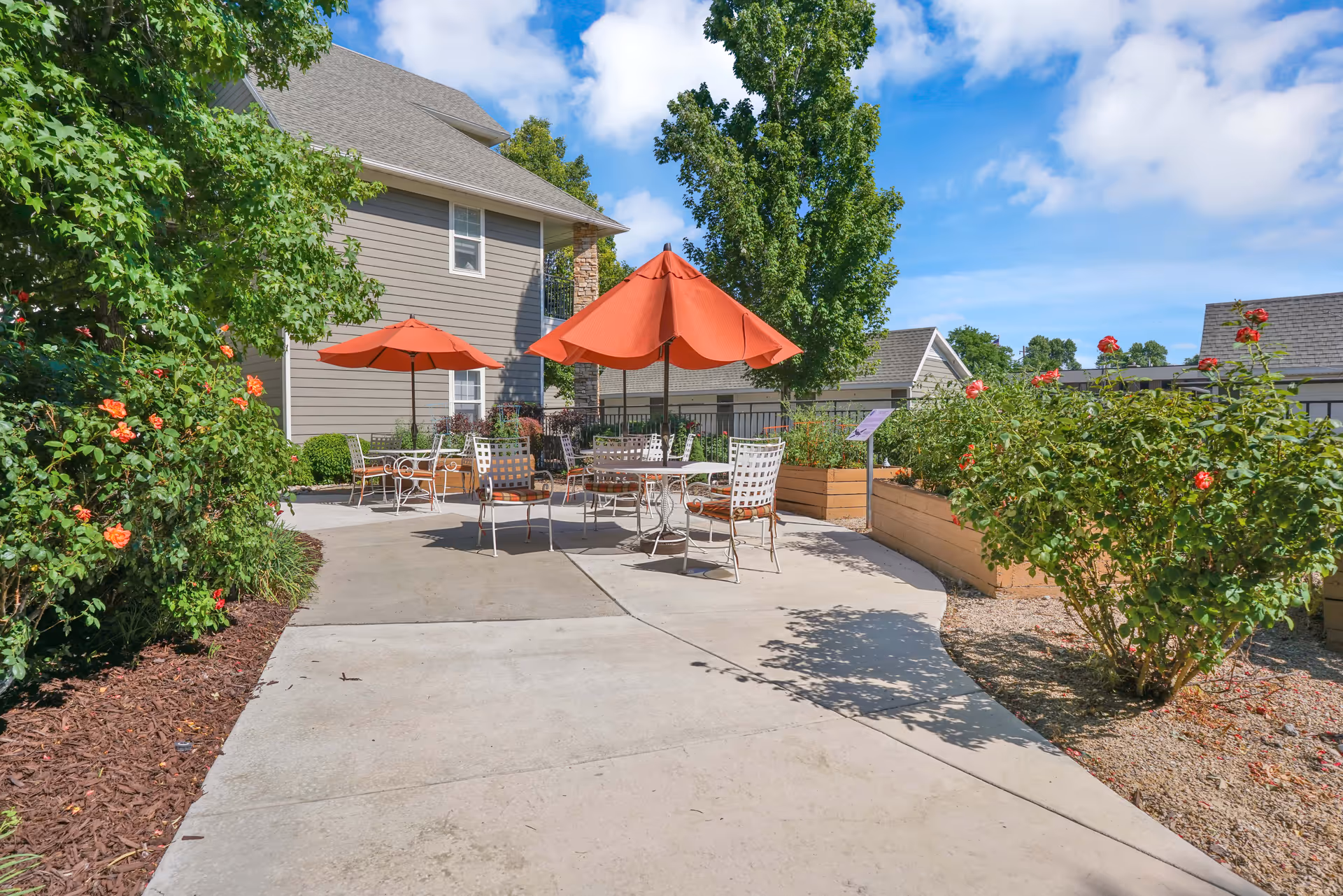 Outdoor patio area at The Wellington with several metal tables and chairs, each shaded by orange umbrellas. The patio is surrounded by green bushes and flowering plants, with a building and blue sky in the background.