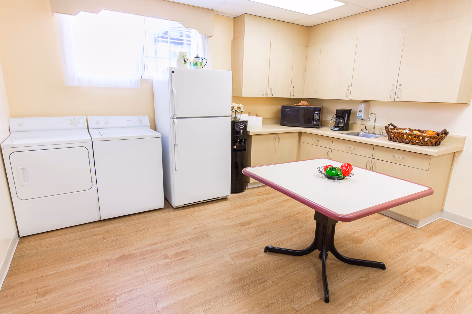 A bright room with light wood flooring and beige walls featuring a white refrigerator, a white washing machine and dryer, light wood cabinets, a microwave, a coffee maker, a sink, and a table with a bowl of decorative peppers in the center.