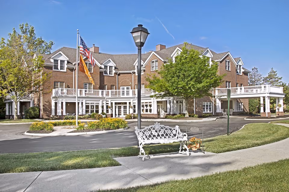 Exterior view of a large brick senior living facility building with multiple windows and a covered entrance. There are two flagpoles with an American flag and another flag, a white decorative bench on a sidewalk, a streetlamp, and well-maintained landscaping with green grass, trees, and flowers under a clear blue sky.