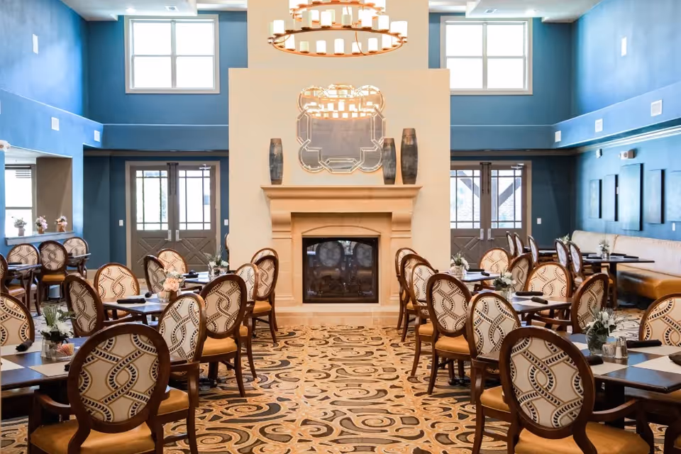 A spacious dining room with multiple tables and patterned chairs arranged neatly. The room features a central fireplace with a decorative mirror and vases on the mantel. The walls are painted blue with large windows letting in natural light, and there is a patterned carpet on the floor.