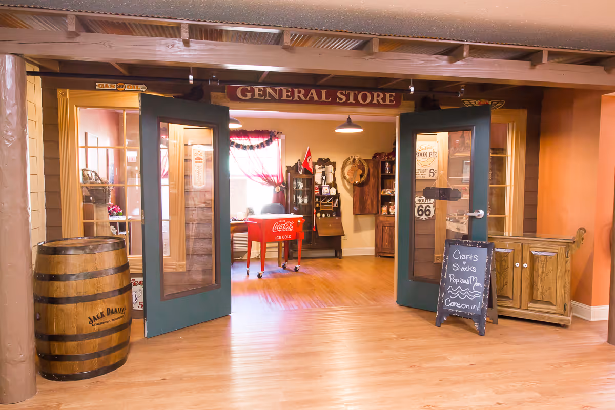 Entrance to a general store area inside a facility with double green doors open. Inside, there is a red Coca-Cola cooler, wooden furniture, and various vintage-style decorations including a Route 66 sign and a Jack Daniel's barrel. A chalkboard sign outside the door advertises crafts, snacks, popcorn, and more. The floor is wooden and the walls are painted warm tones.
