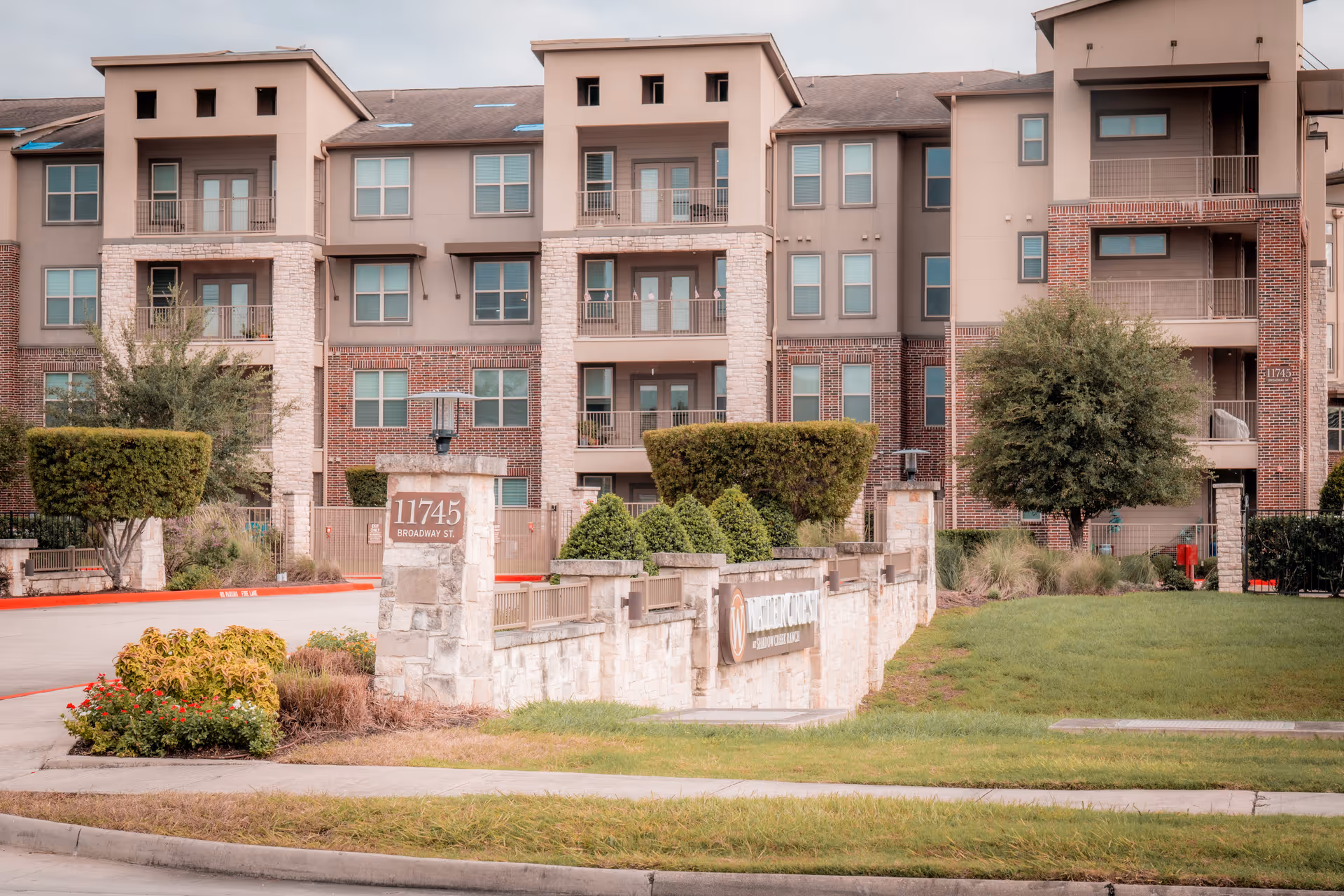 Exterior view of a multi-story residential building with balconies and numerous windows. In front of the building is a stone sign displaying the address 11745 Broadway St. and a sign for Watercrest at Shadow Creek Ranch, surrounded by neatly trimmed bushes and landscaping.