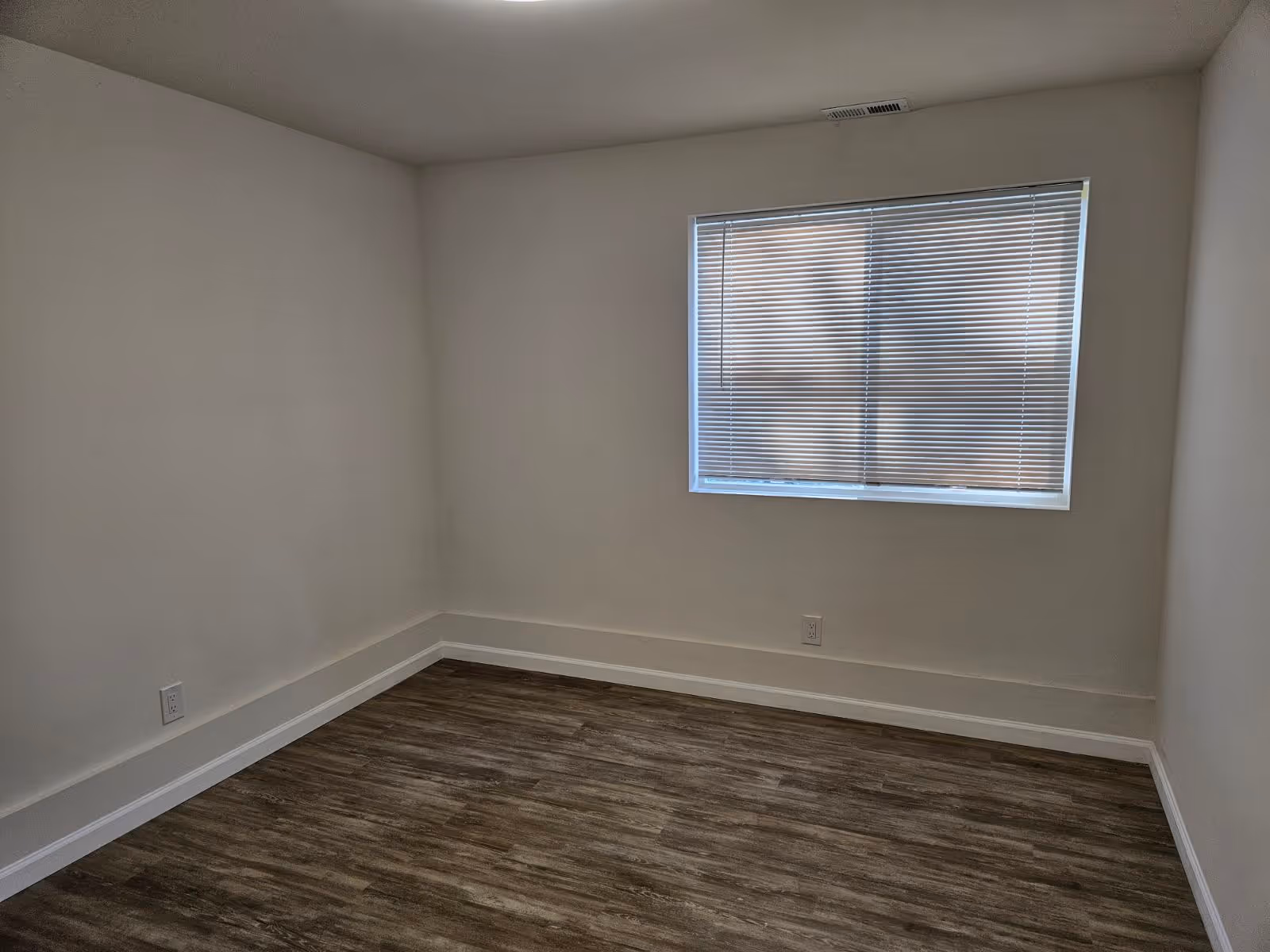 Empty room with white walls, a window with closed blinds, and wood-patterned flooring.