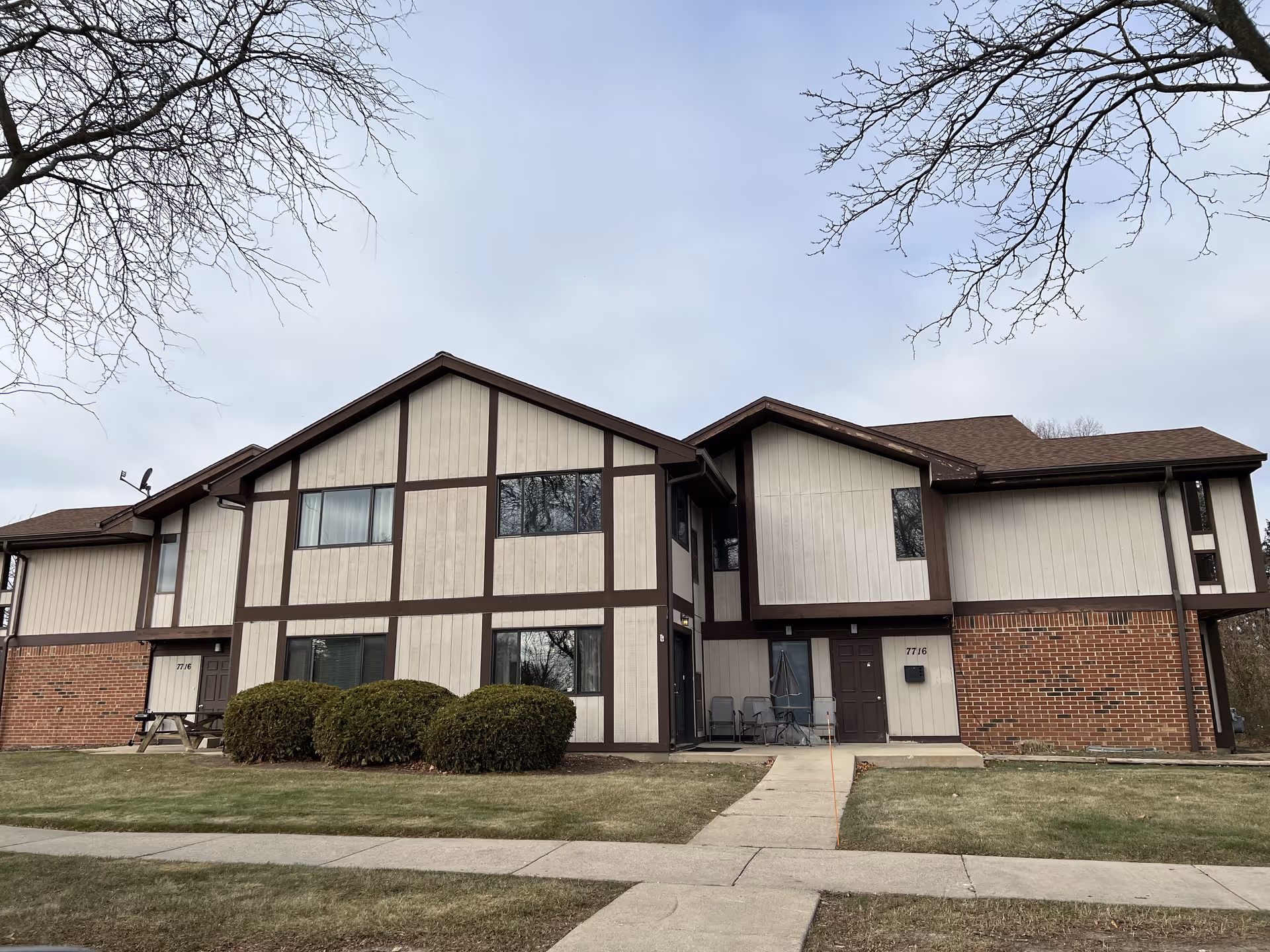 Exterior view of a two-story residential building with beige and brown paneling and brick accents. There are several windows, a few bushes in front, a picnic table on the left side, and a small patio area with chairs and an umbrella on the right side. The sky is overcast and there are leafless tree branches visible at the top edges of the image.
