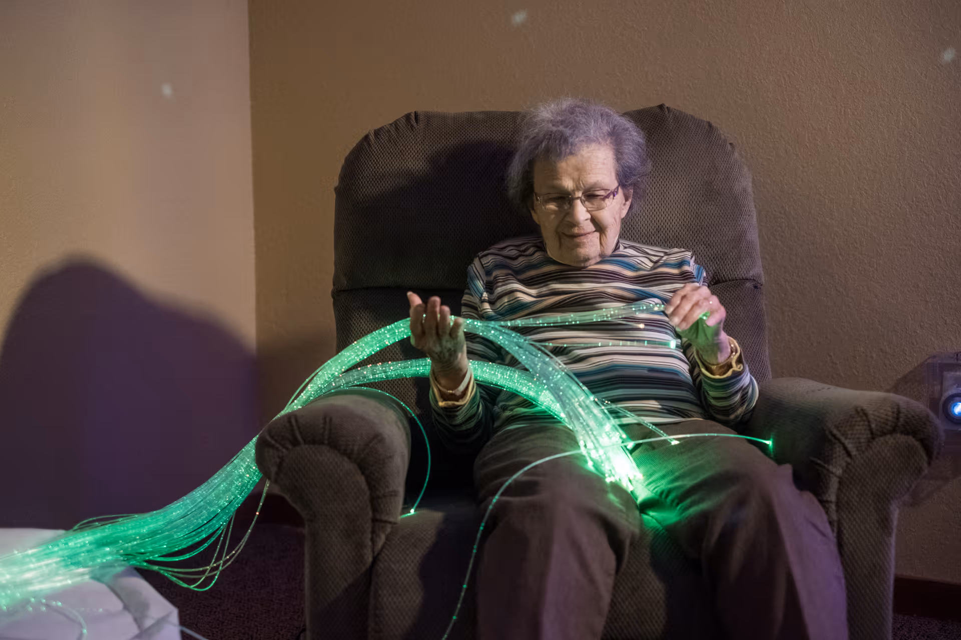 An elderly woman sitting in a cushioned armchair, holding and looking at glowing green fiber optic strands in a dimly lit room.