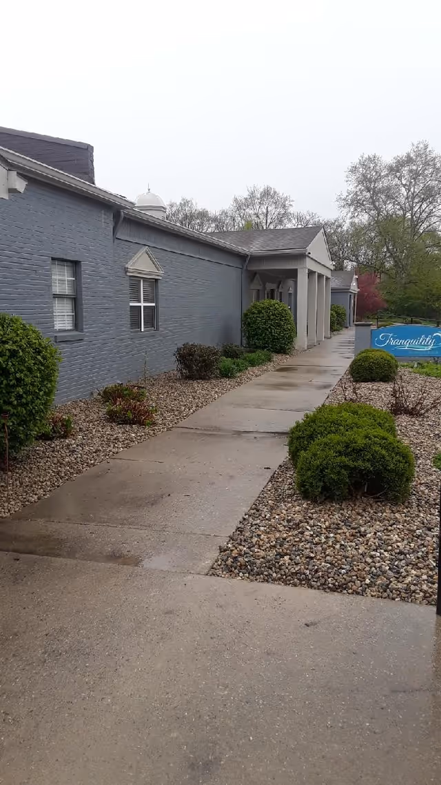 Sidewalk leading to the entrance of a single-story building with gray brick walls and white trim. The walkway is bordered by small bushes and landscaping rocks. A blue sign with the word 'Tranquility' is visible near the entrance. Trees and greenery are in the background under an overcast sky.