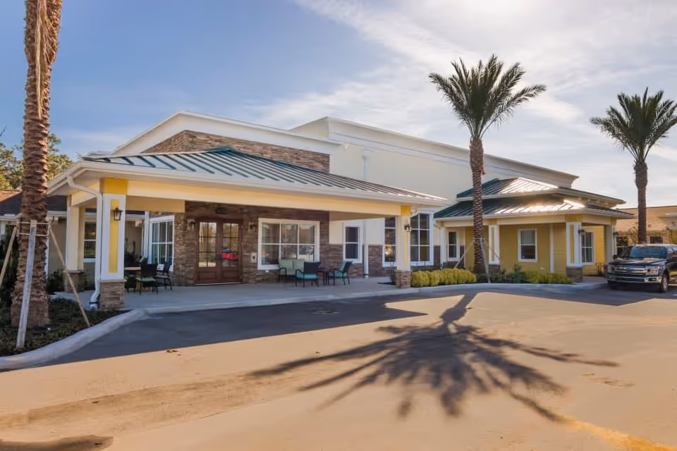 Exterior view of CERTUS Premier Memory Care Living facility showing the entrance with a covered drop-off area, palm trees, and a parked vehicle on a sunny day.