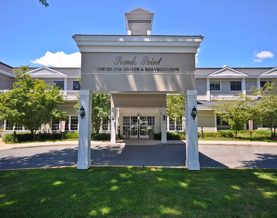 Front entrance of Sands Point Center for Health & Rehabilitation, a two-story building with white columns and a covered driveway, surrounded by green trees and grass under a clear blue sky.