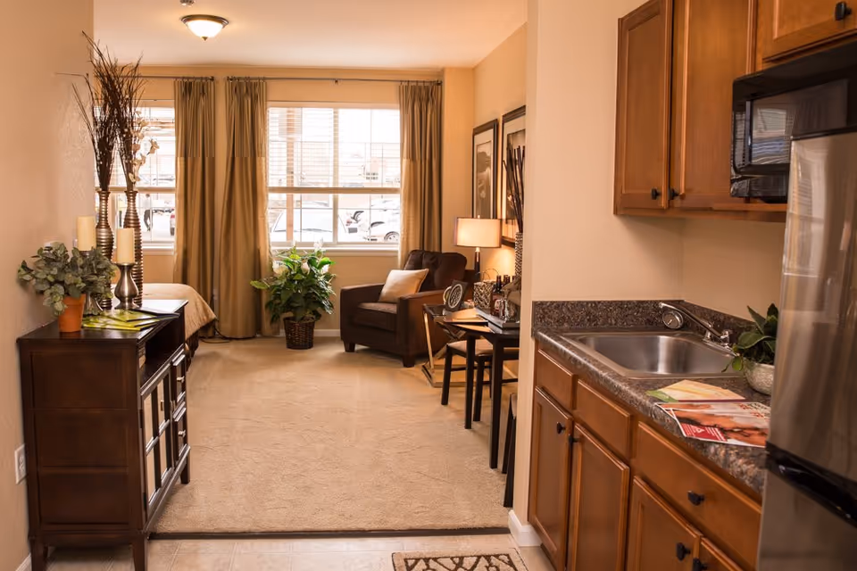 Interior view of a senior living facility apartment at Traditions of Hilliard showing a small kitchen area with wooden cabinets, a sink, and a refrigerator on the right. The living area includes a brown armchair, a small table with a lamp, framed pictures on the wall, and a window with beige curtains letting in natural light. There is also a wooden sideboard with decorative plants and candles on the left.