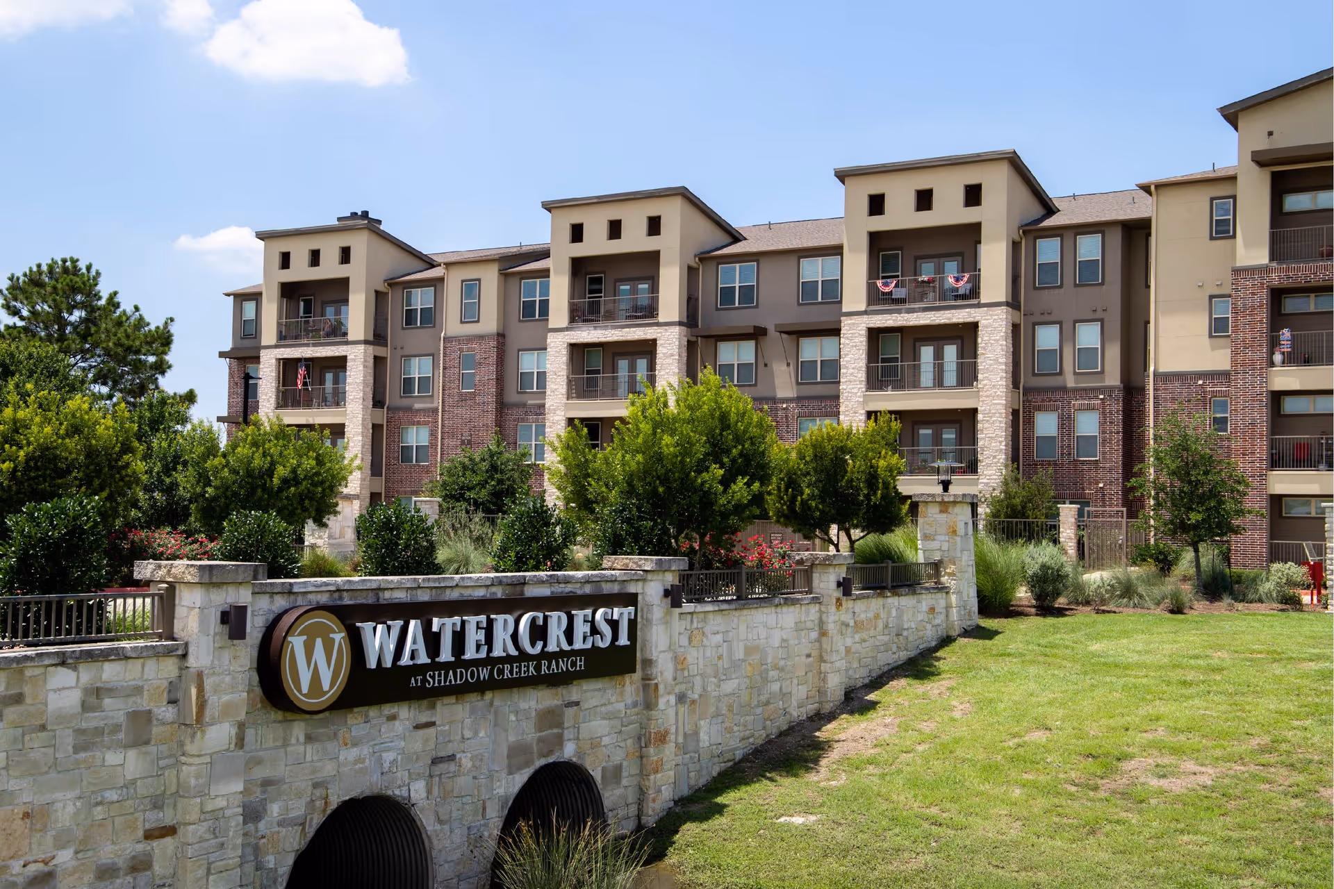 Exterior view of a multi-story senior living facility building with balconies, surrounded by greenery and a stone wall with a sign that reads 'Watercrest at Shadow Creek Ranch'.