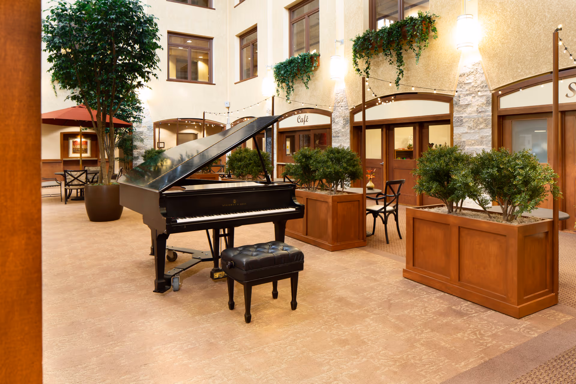 Indoor common area with a black grand piano and matching piano bench in the center. The space features potted plants, wooden planters with greenery, tables and chairs, and warm lighting. There are windows and doors along the walls, with a sign above one door reading 'Café'.