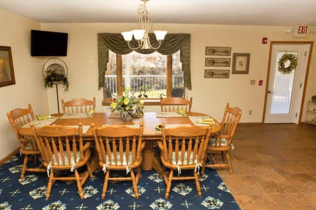 A dining room with a large wooden table surrounded by eight wooden chairs with cushions. The table is set with placemats and napkins, and a floral centerpiece is in the middle. There is a window with green curtains behind the table, a wall-mounted TV to the left, and a door with a wreath on it to the right. The floor is a combination of tile and a blue patterned rug under the table.