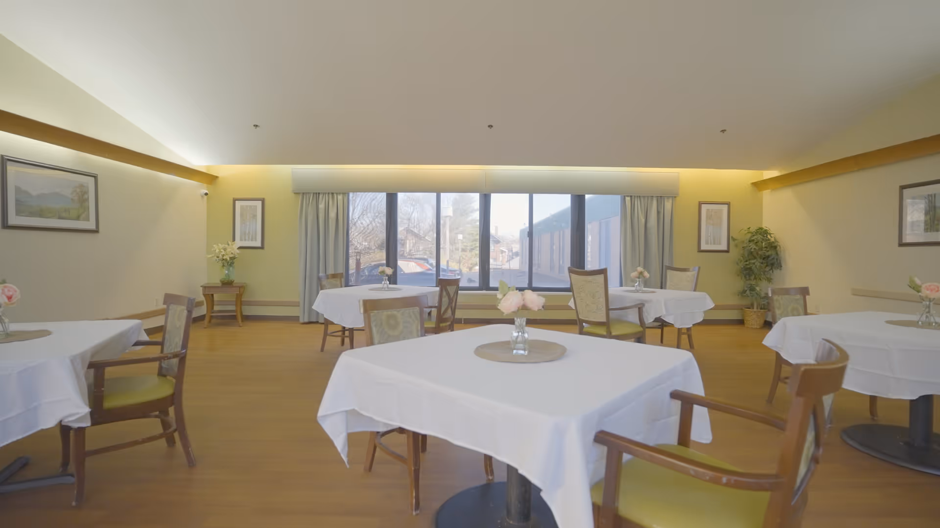 A bright dining room with several tables covered in white tablecloths, each adorned with a small vase of flowers. Wooden chairs with green cushions surround the tables. Large windows at the far end let in natural light, and framed artwork and plants decorate the walls.