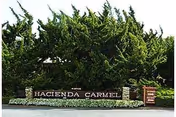 Entrance sign for Hacienda Carmel Community surrounded by green trees and bushes, with a stone base and a smaller sign to the right.