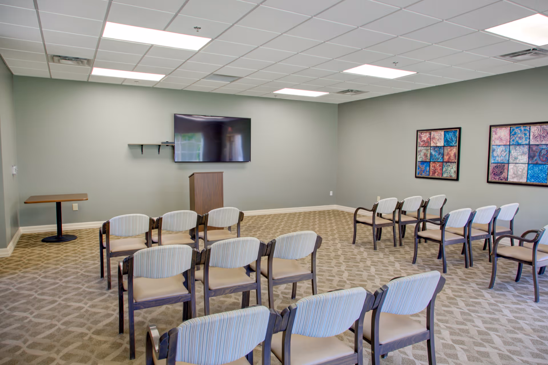 A conference or meeting room with rows of chairs facing a wall-mounted flat screen TV and a wooden podium. The room has light green walls, patterned carpet, two colorful framed artworks on one wall, and a small table in the corner.