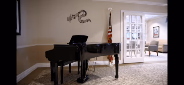 A black grand piano in a carpeted room with beige walls and white trim. Behind the piano is an American flag and a wall decoration featuring musical notes. To the right, there is an open doorway with glass-paneled double doors leading to a seating area with chairs and framed artwork on the wall.