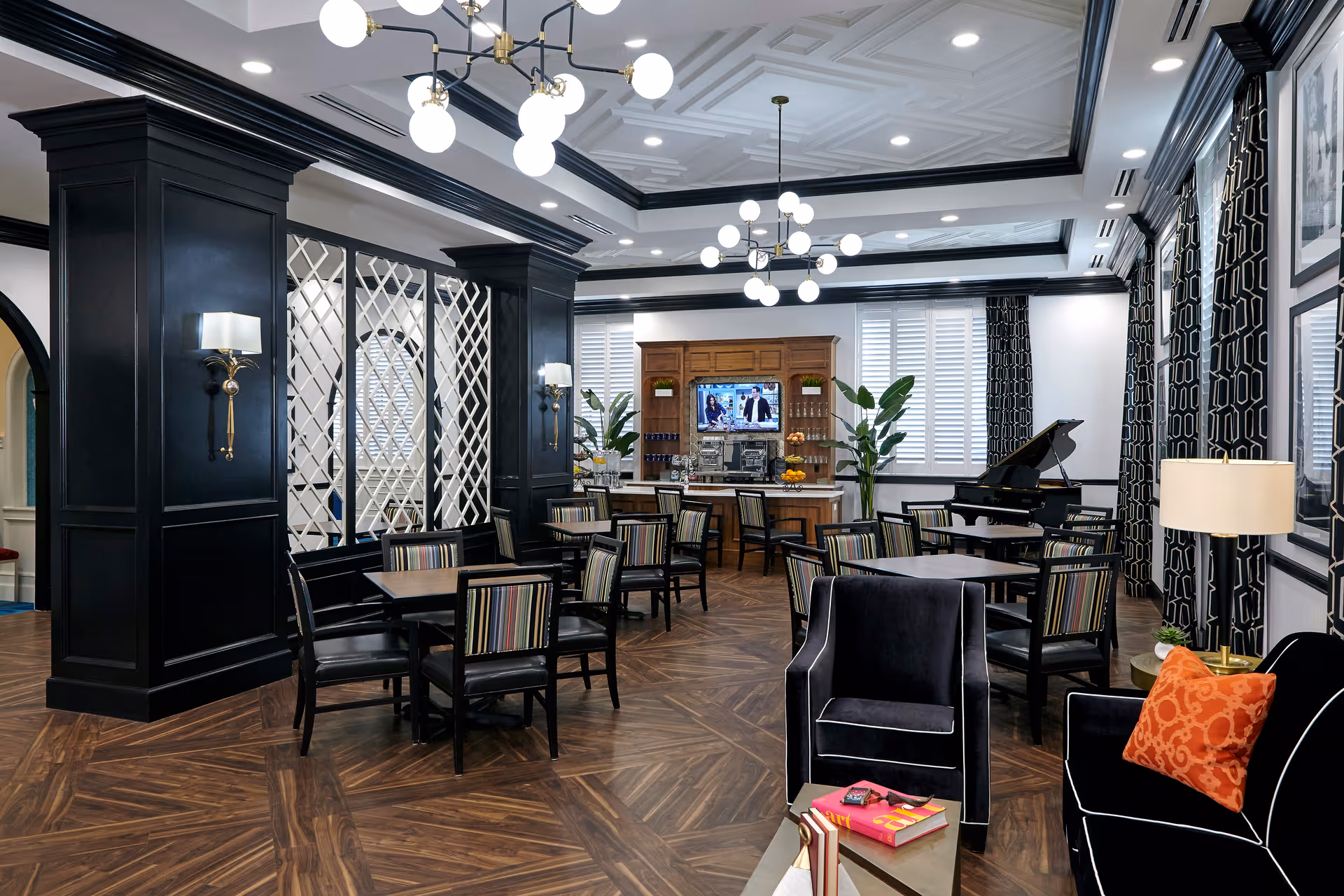 A stylish and elegant common area in an assisted living facility featuring multiple tables with striped chairs, a black grand piano, a wooden cabinet with a television mounted above it, and large windows with white shutters and black patterned curtains. The room has dark wood flooring, black paneled columns, modern chandeliers, and comfortable seating including a black armchair with an orange pillow.