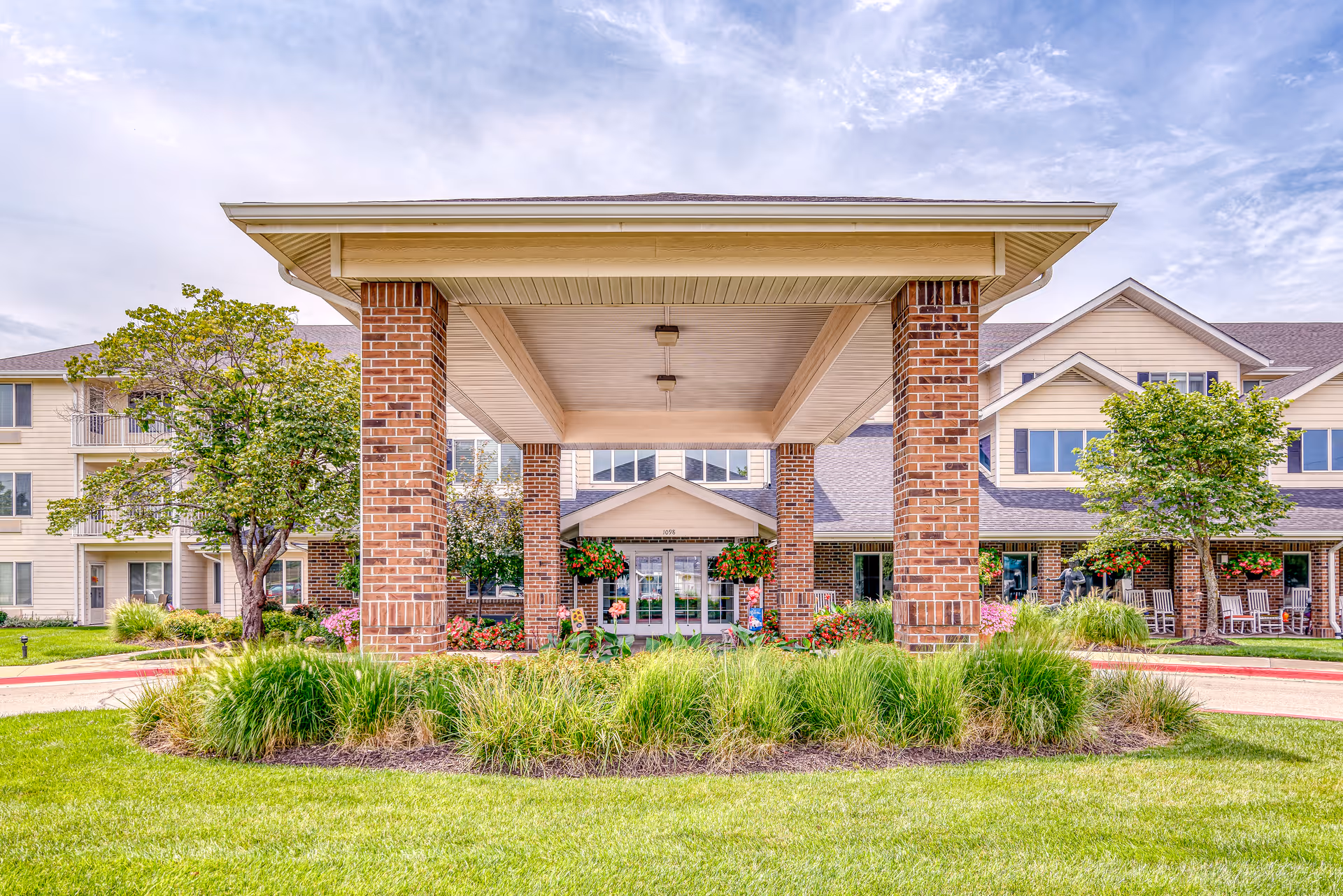 Front exterior view of Solstice Senior Living at Lee's Summit showing a covered entrance supported by brick pillars, surrounded by green grass, bushes, and trees under a partly cloudy sky.