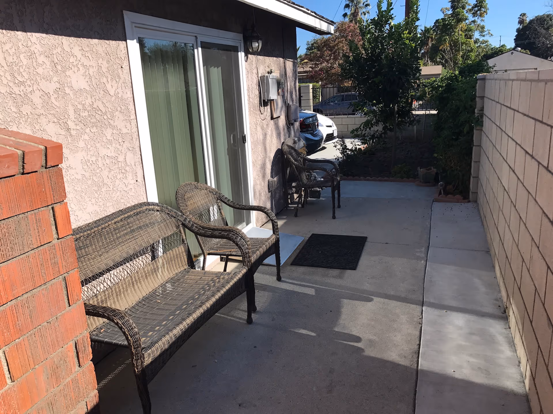 Outdoor patio area with two wicker chairs and a wicker bench placed on a concrete floor next to a sliding glass door. There is a brick wall on the left and a beige stucco wall on the right, with some greenery and parked cars visible in the background.