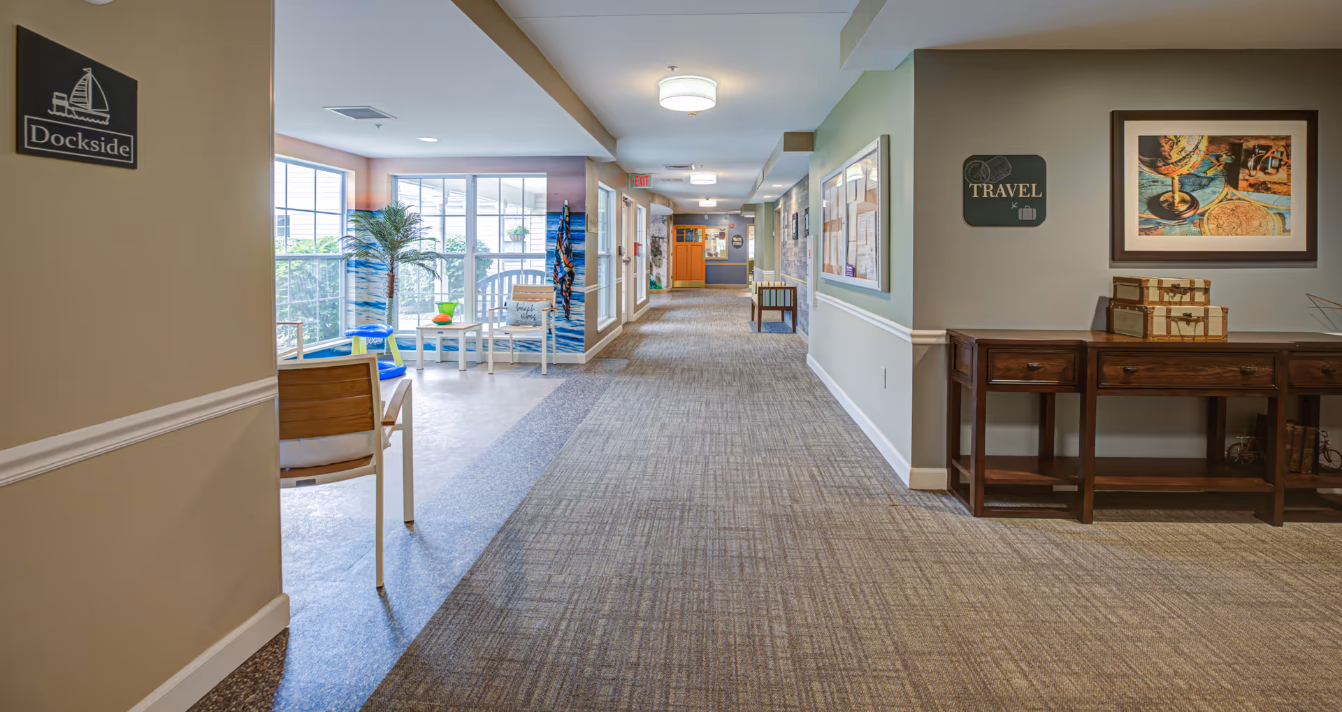 A well-lit hallway in a senior living facility with carpeted flooring and beige walls. On the left side, there is a seating area with white chairs and a small table near large windows letting in natural light. The wall on the left has a sign that reads 'Dockside'. On the right side, there is a wooden console table with decorative items and a framed picture above it, along with a sign that says 'Travel'. The hallway extends into the distance with more seating and bulletin boards visible.