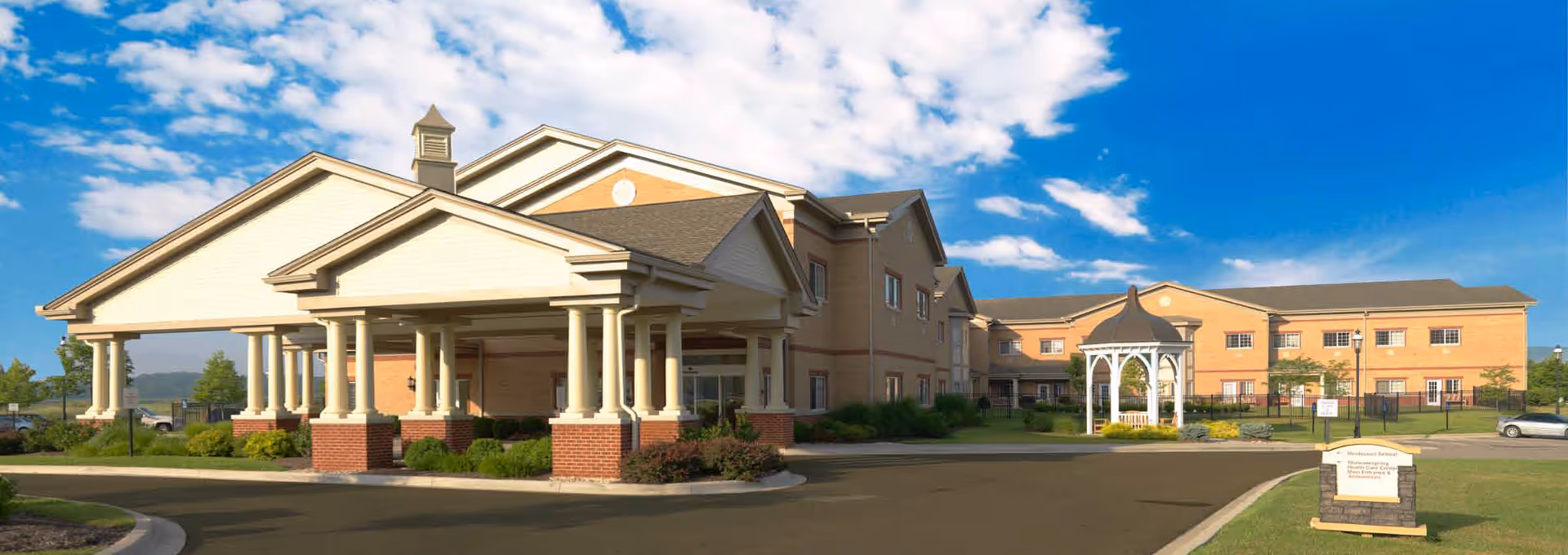 Exterior view of Shawneespring Skilled Nursing Facility showing a large building with a covered entrance supported by columns, a gazebo, landscaped greenery, and a clear blue sky with some clouds.