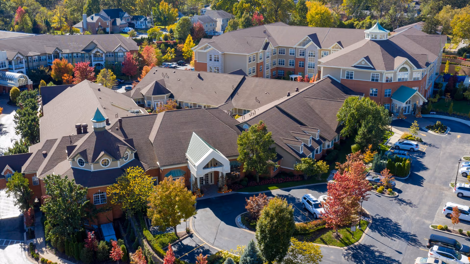 Aerial view of McKnight Place Extended Care facility showing a large multi-wing building with a mix of brick and siding exterior, surrounded by trees with autumn foliage and a parking lot with several cars.