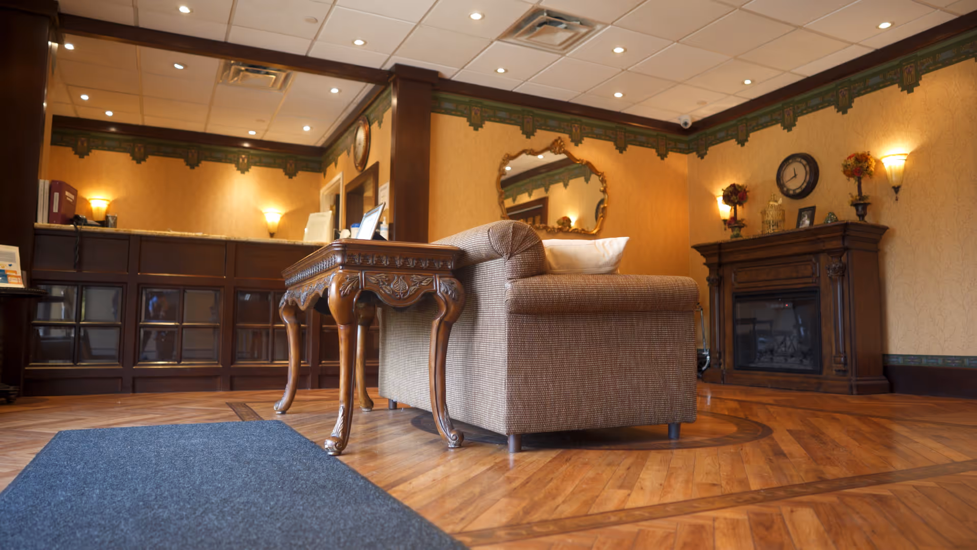 A cozy interior room with wooden flooring and warm yellow walls featuring decorative trim. There is a brown upholstered armchair next to an ornate wooden side table. In the background, there is a wooden reception desk with a granite countertop, a wall clock, a decorative mirror, and a wooden fireplace mantel with small decorative items and flowers on top.