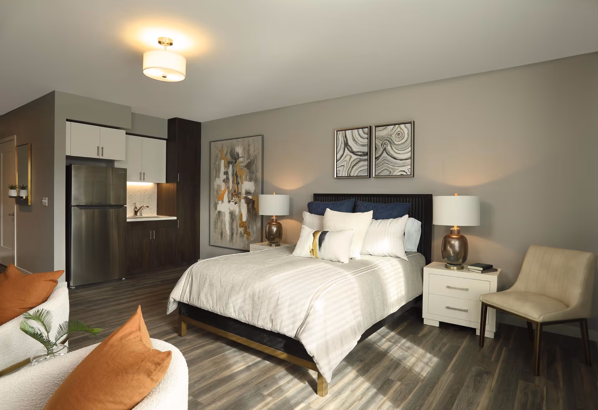 A modern studio apartment with a bed in the center, flanked by two nightstands with lamps. The bed has multiple pillows and a beige bedspread. To the left is a small kitchenette with a stainless steel refrigerator, sink, and cabinets. There is a beige chair next to the right nightstand and two abstract paintings on the wall above the bed. The floor is wood, and the walls are painted light gray. Two orange pillows are visible on a white chair in the foreground.