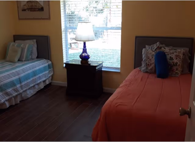 A bedroom with two single beds separated by a dark wooden nightstand with a blue lamp on it. The bed on the left has a striped bedspread with pillows, and the bed on the right has a solid coral bedspread with multiple pillows. A window with blinds is centered on the wall behind the nightstand, letting in natural light. The room has wooden flooring and yellow walls.