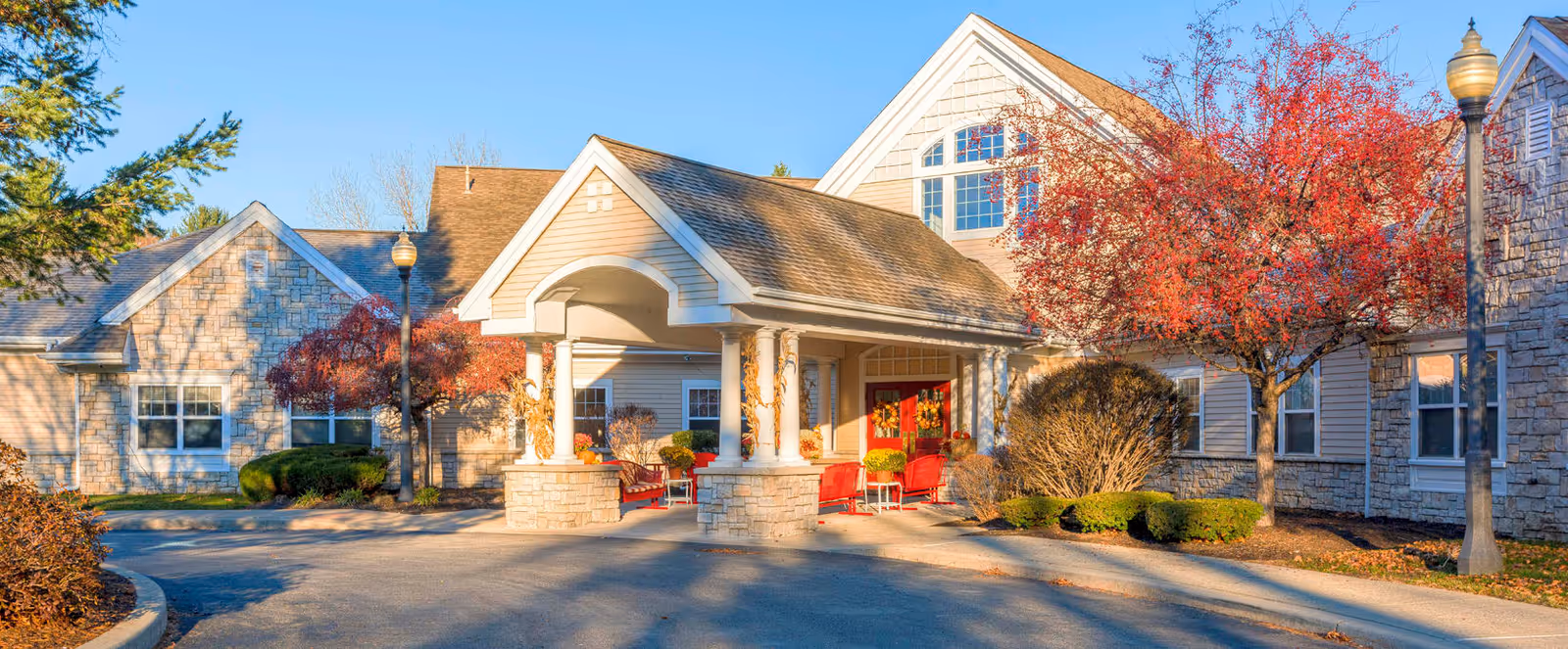 Exterior view of a senior living facility building with a covered entrance supported by columns. The building features stone and siding walls, multiple windows, and a peaked roof. There are red-leaved trees and well-maintained shrubs around the entrance area, with a paved driveway leading up to the entrance.