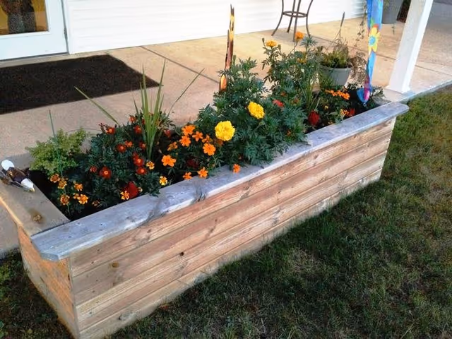 A wooden rectangular planter box filled with various blooming flowers including yellow and orange marigolds, placed outdoors on grass near a building entrance with a concrete porch and a door.