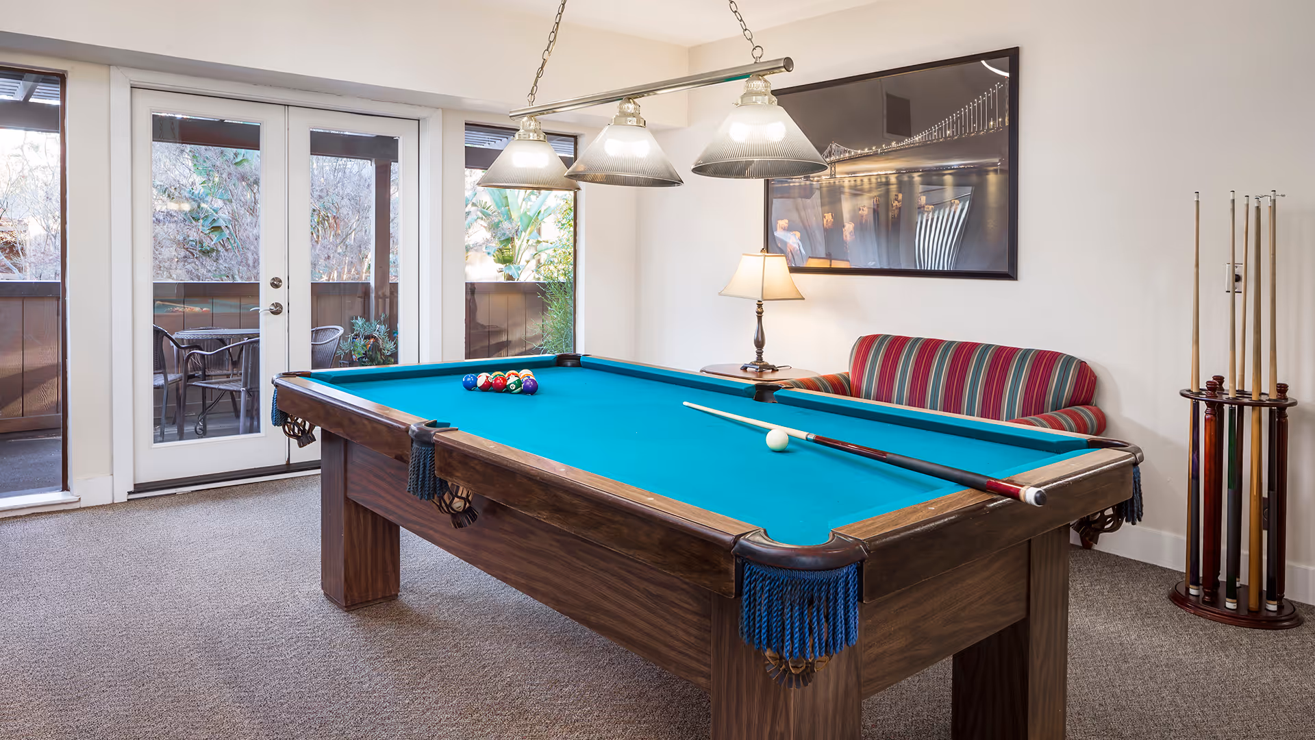A well-lit room with a pool table in the center, featuring a blue felt surface and a set of billiard balls arranged for a game. Above the pool table, three hanging pendant lights provide illumination. To the right, there is a striped upholstered couch with a side table and a lamp. A framed picture hangs on the wall above the couch. In the background, glass doors lead to an outdoor patio area with chairs and a table. A rack holding pool cues stands near the wall on the right side.
