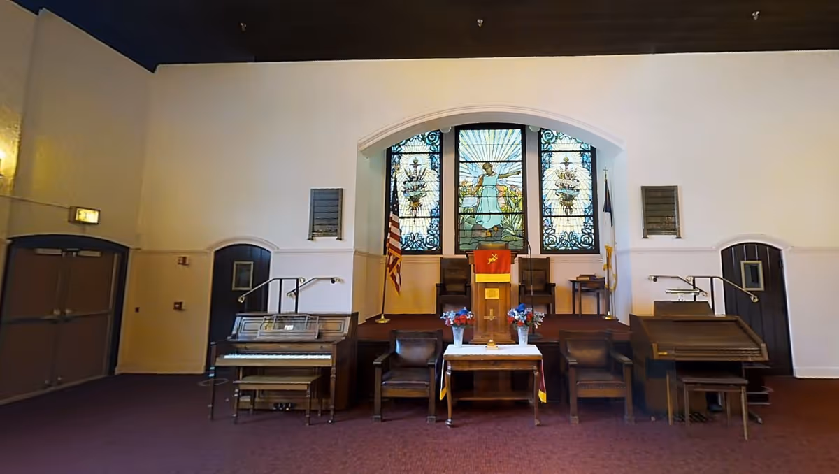 Interior view of a chapel or worship room with a stained glass window depicting an angel behind a podium draped with a red cloth. There are two flags on either side of the podium, a piano on the left, a lectern on the right, and several chairs arranged around a small table with flower arrangements.
