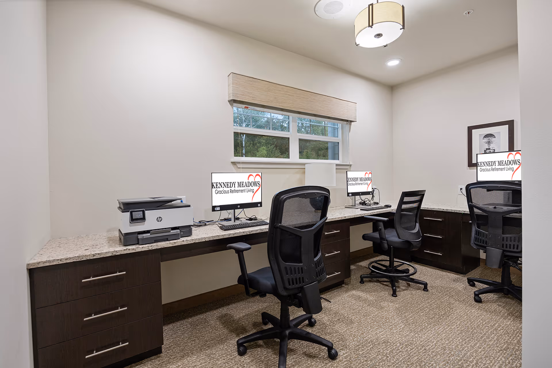 A small office room with three black mesh office chairs and three computer workstations on a long L-shaped desk. Each computer screen displays the logo and text 'KENNEDY MEADOWS Gracious Retirement Living'. There is a printer on the left side of the desk and a window with a beige blind above the desk. The room has beige walls and carpeted floor with a ceiling light fixture.