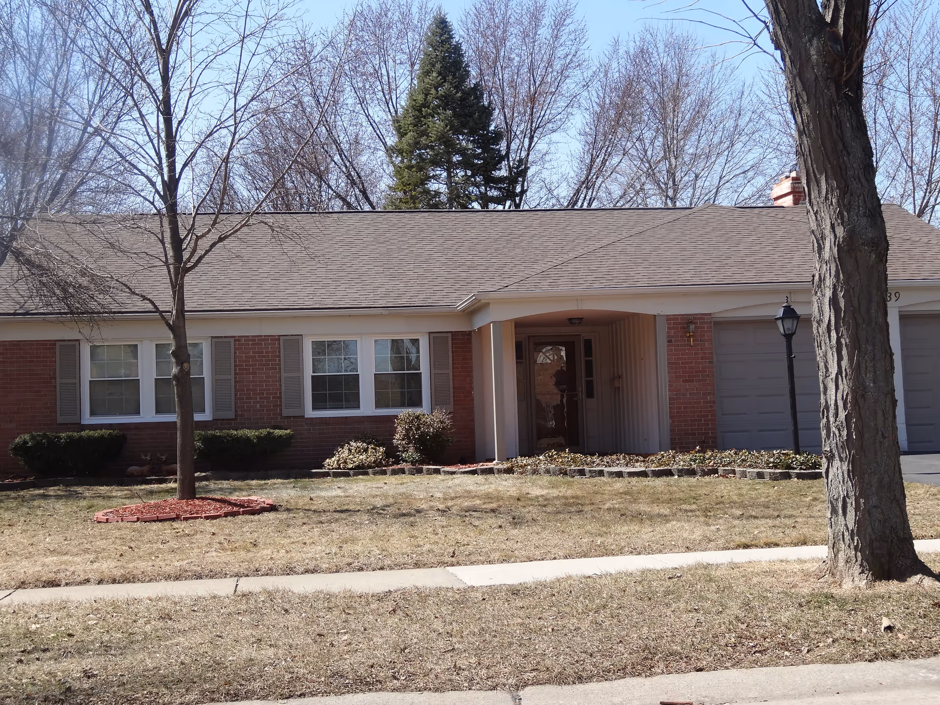 Single-story brick house with a front porch, two large windows with shutters, a garage with two doors, a lamp post, and leafless trees in the front yard during winter or early spring.