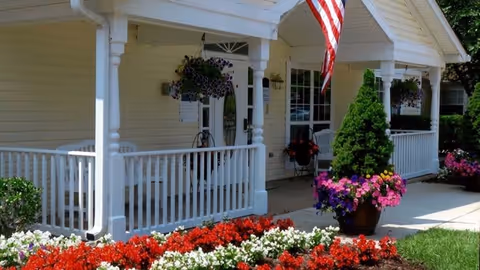 Front porch area of a light yellow building with white railings and columns, decorated with hanging flower baskets and potted plants. An American flag is displayed near the entrance, and vibrant red, white, and purple flowers are planted in the garden bed in front.