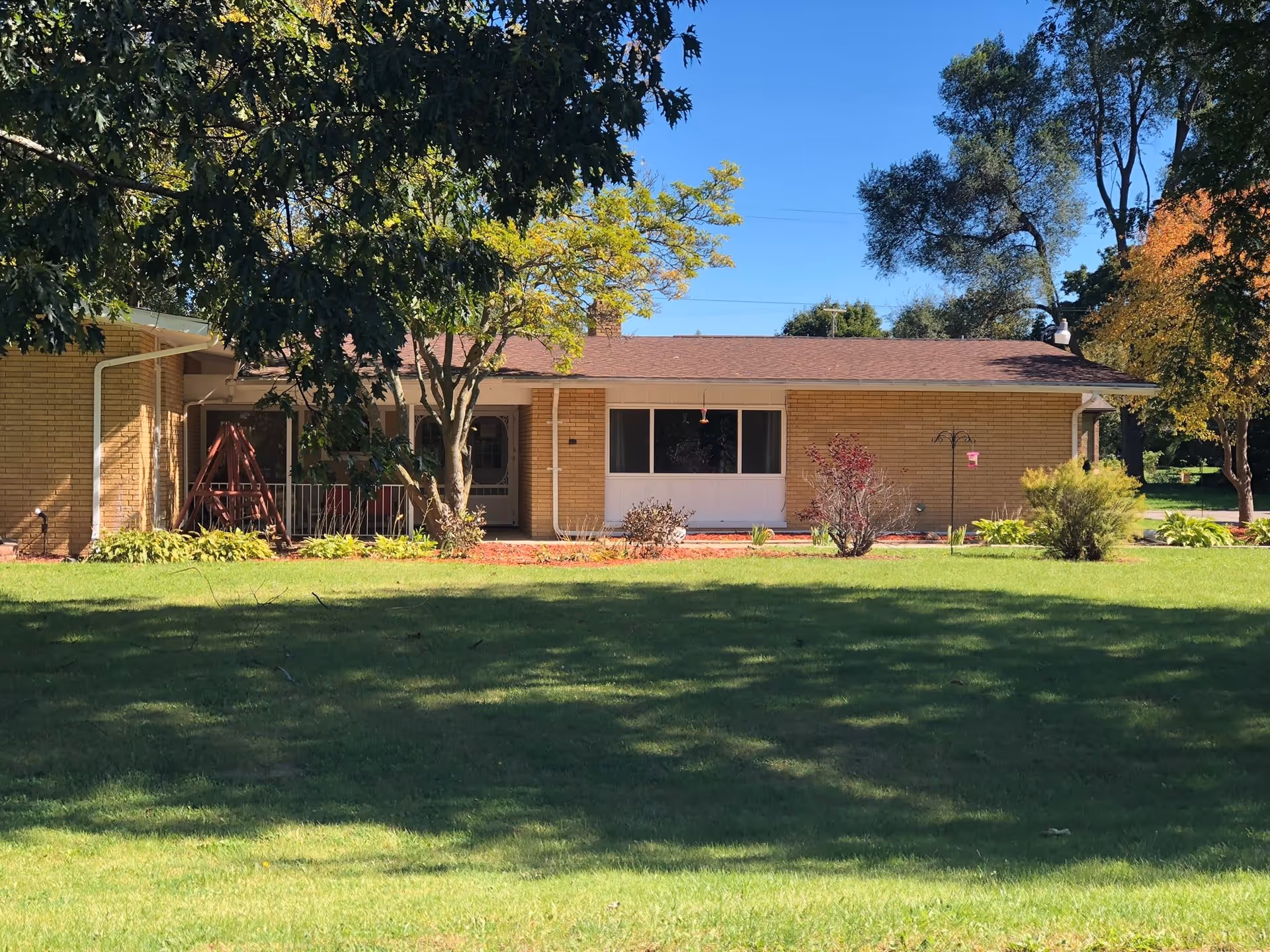 Single-story brick building with a brown roof surrounded by green grass and trees under a clear blue sky. There is a porch area with a swing and some bushes and plants in front of the building.