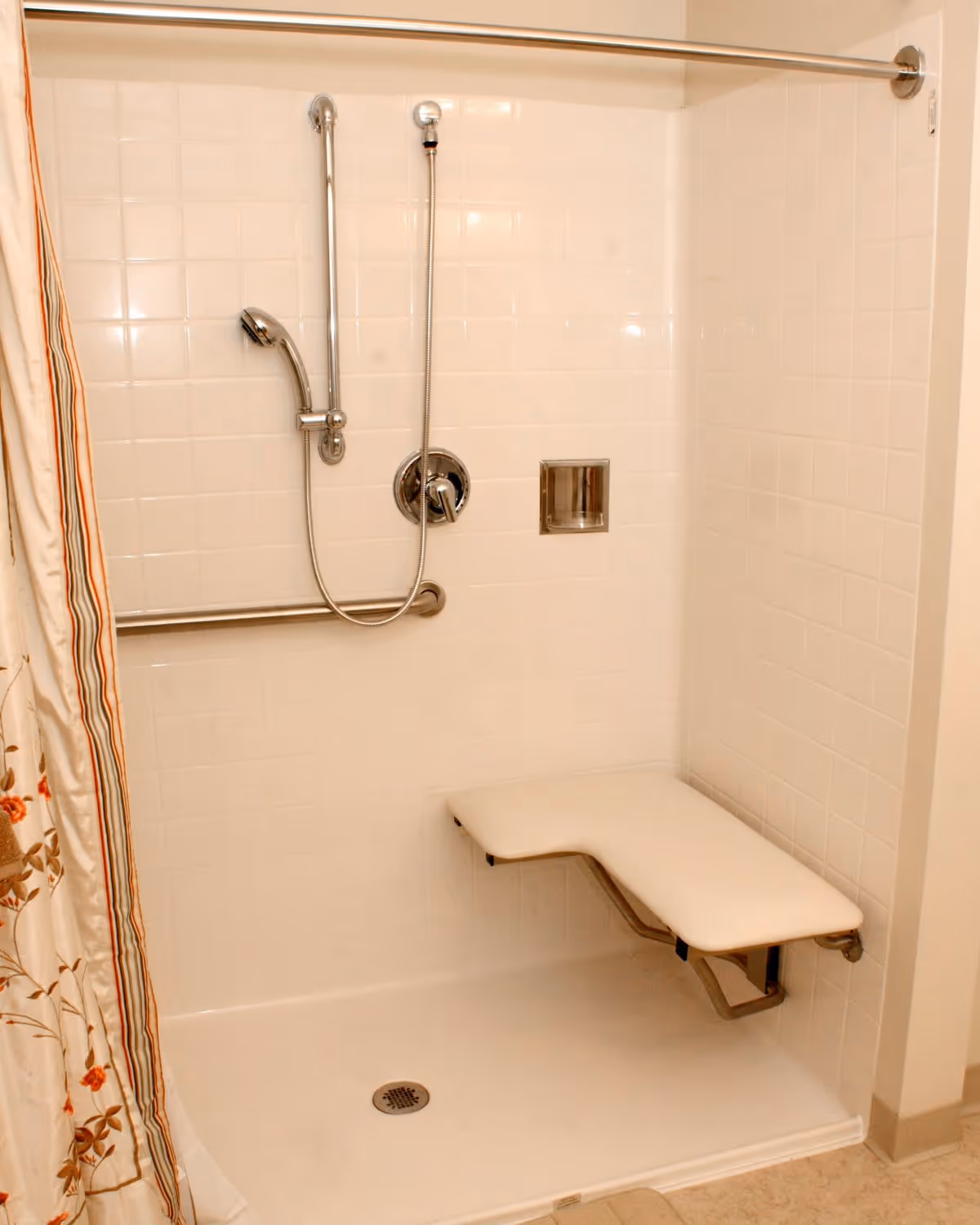 A white tiled shower area with a fold-down seat, a handheld showerhead attached to a vertical grab bar, a horizontal grab bar, and a floral-patterned shower curtain partially visible on the left side.
