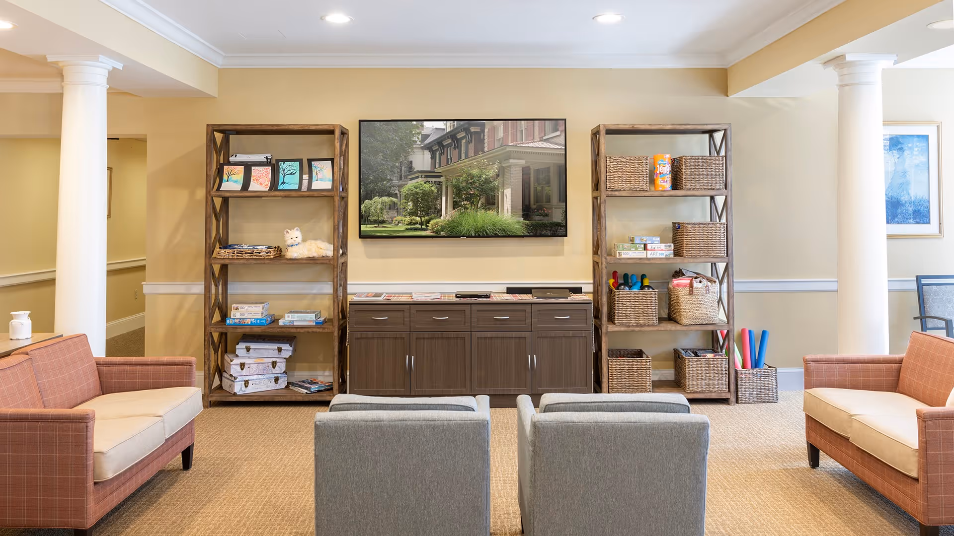 A bright and cozy living room area with two orange sofas facing two gray armchairs. Between the sofas are two tall wooden shelves filled with baskets, books, and decorative items. A large flat-screen TV is mounted on the wall above a dark wooden cabinet. The room features beige walls, white columns, and a framed picture on the right wall.