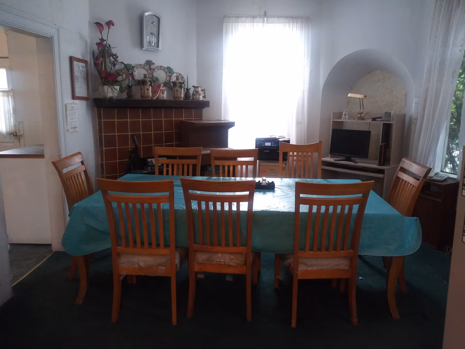 A dining room with a rectangular table covered with a blue tablecloth surrounded by eight wooden chairs. The room has a window with sheer white curtains letting in natural light. There is a fireplace with decorative plates and plants on the mantel, a clock above it, and a small desk with a computer and lamp in an alcove to the right.