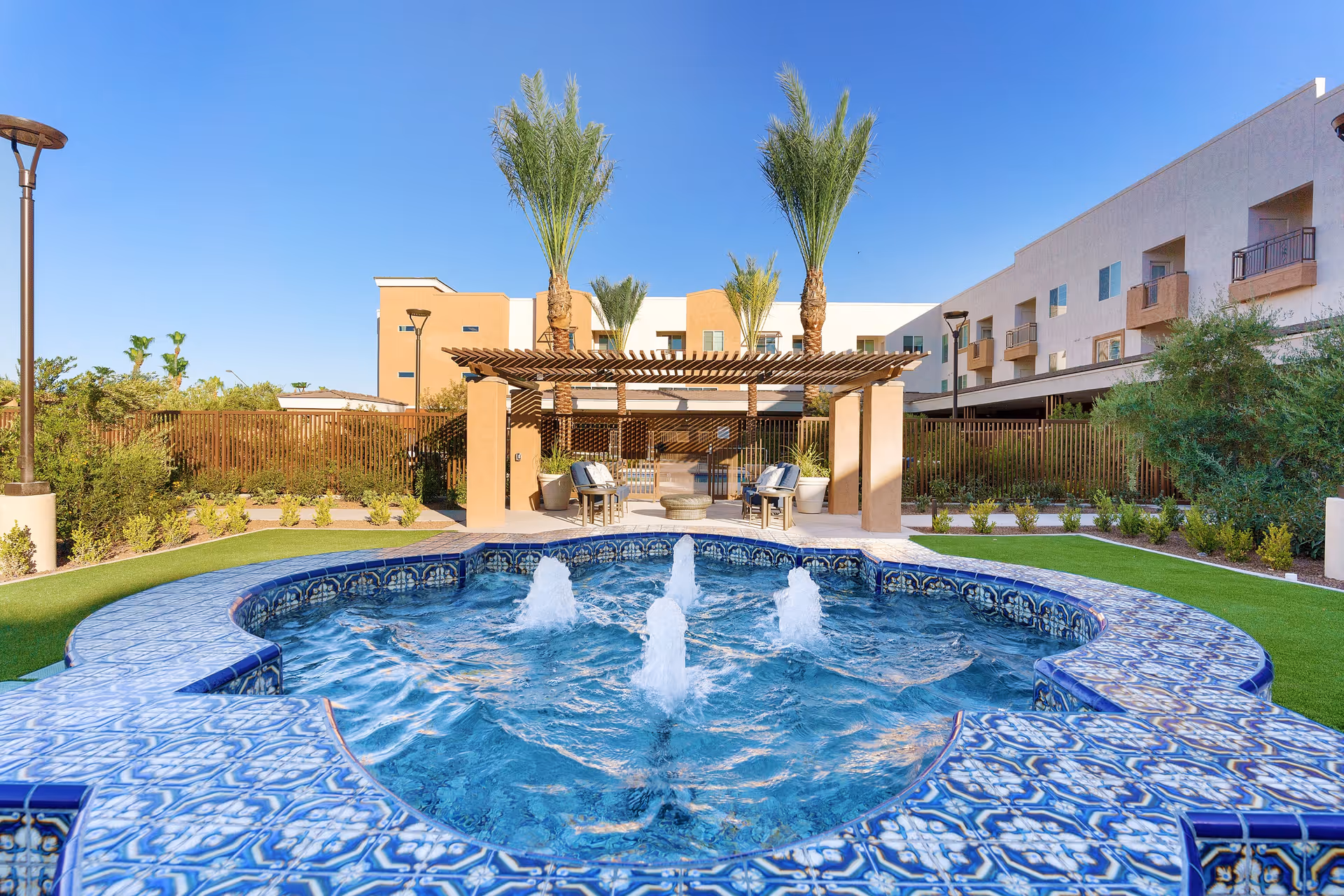 Outdoor area at Robson Reserve at PebbleCreek featuring a decorative blue-tiled fountain with water jets in the foreground, surrounded by green landscaping and palm trees. In the background, there is a pergola with seating and a multi-story building under a clear blue sky.