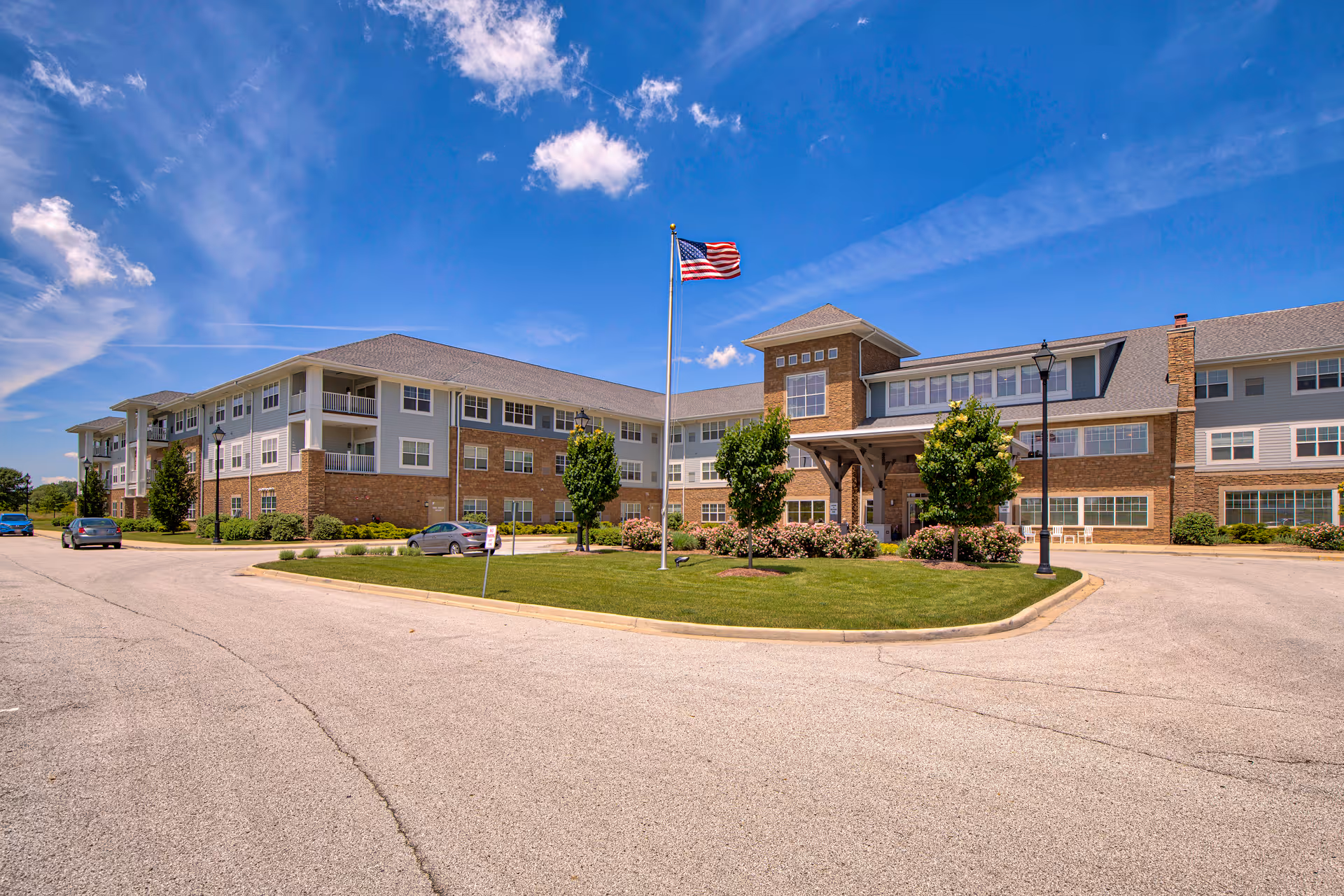 Exterior view of a large, three-story senior living facility building with a combination of brick and light-colored siding. There is a circular driveway with a grassy island in the center featuring an American flag on a flagpole and several small trees and shrubs. The sky is clear with a few clouds.