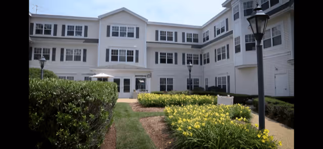Exterior view of a multi-story white senior living facility building with multiple windows, surrounded by landscaped gardens with green bushes and yellow flowers, and a paved walkway with black lamp posts.