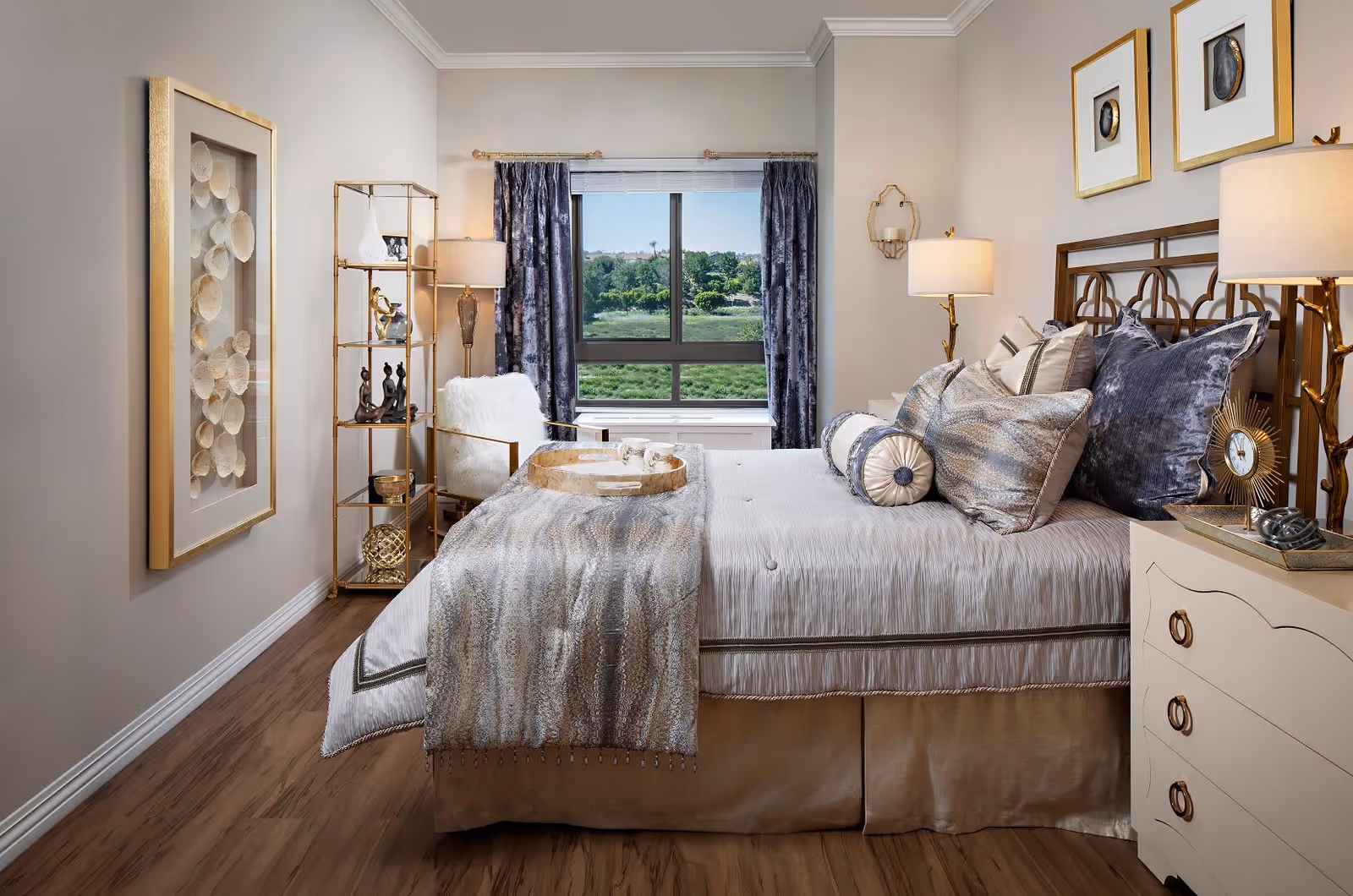 A well-decorated bedroom with a large bed adorned with multiple pillows and a throw blanket. There is a window with dark curtains showing greenery outside. The room features a white armchair with a fluffy cushion, a gold and glass shelving unit with decorative items, two bedside tables with lamps, and framed artwork on the walls. The floor is wooden.