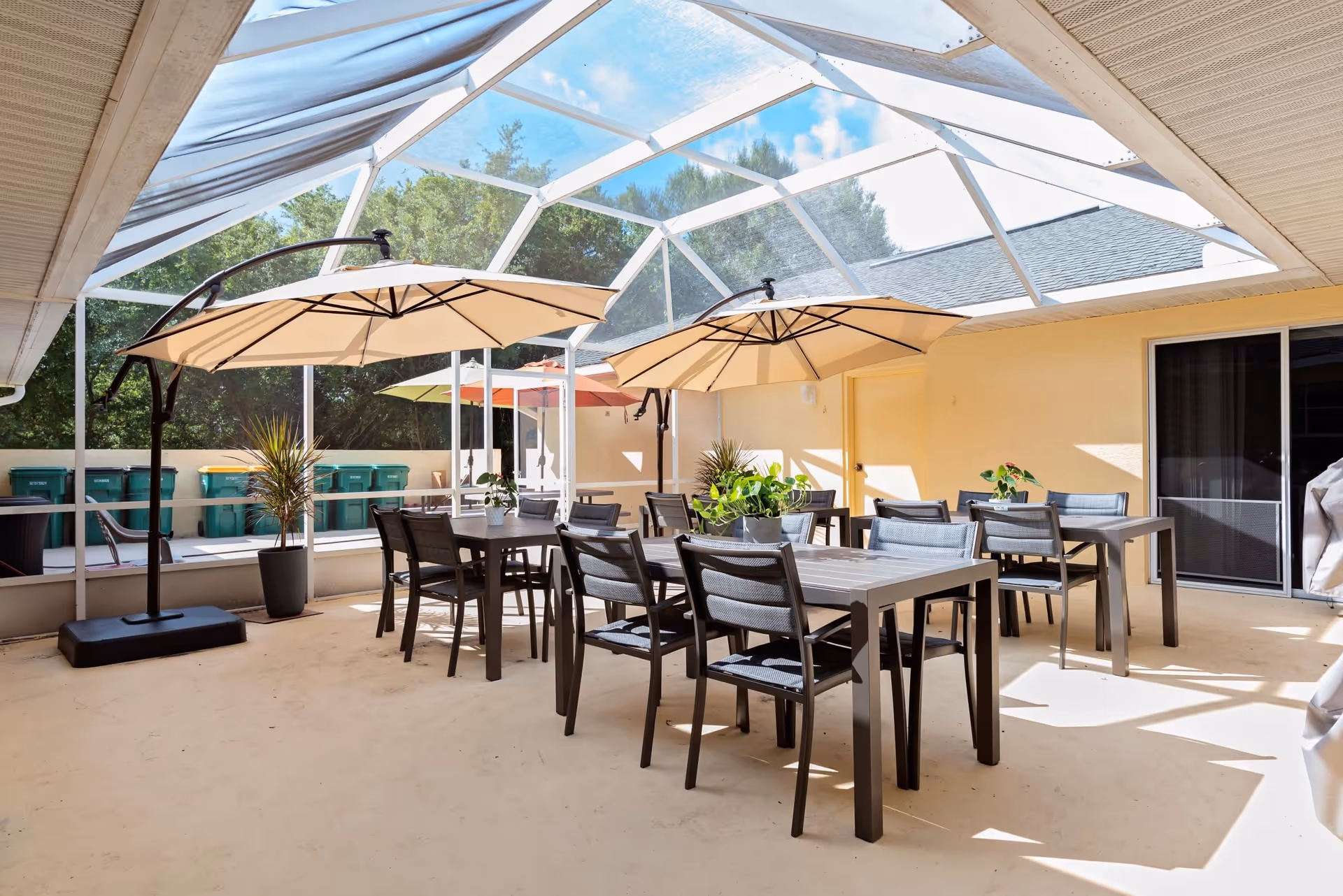 A covered outdoor patio area with multiple tables and chairs arranged for seating. Two large beige umbrellas provide shade over the tables. There are potted plants on the tables and around the patio. The area is enclosed with a transparent roof structure, allowing natural light to come through. Trees and blue sky are visible outside the enclosure.