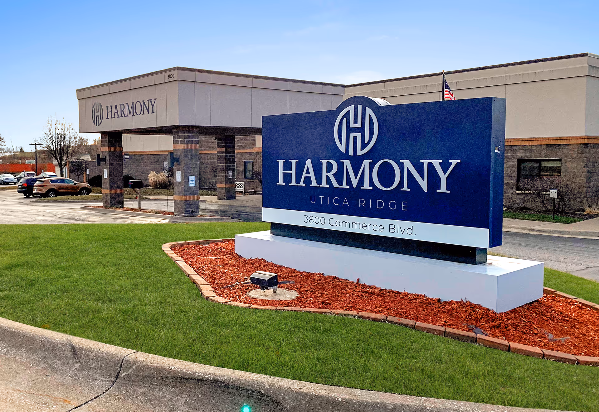 Exterior view of Harmony Utica Ridge facility showing a large blue sign with the facility name and address 3800 Commerce Blvd. The building entrance and parking area are visible in the background under a clear blue sky.