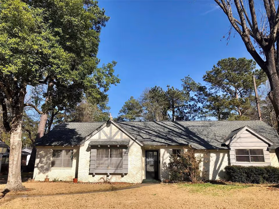 Exterior front view of a single-story residential building with light-colored brick walls and a gray shingled roof. The house has multiple windows with shutters and a central front door. There are large trees and bushes around the house, and the sky is clear and blue.