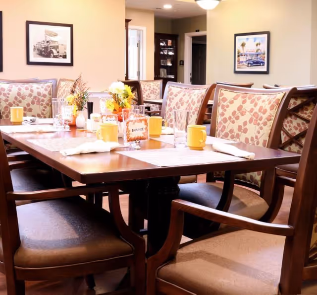 A dining room table set with yellow mugs, glasses, napkins, and small flower arrangements. The chairs around the table have wooden frames and patterned upholstery. The room has warm lighting and framed pictures on the walls.